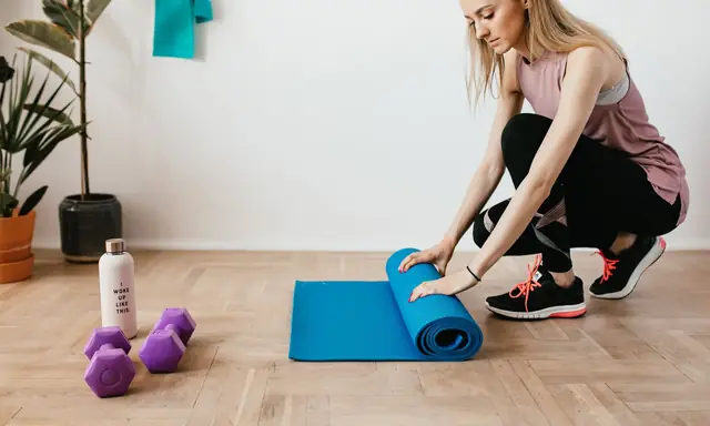 Vrouw in sportkleding rolt thuis een blauwe oefenmat uit op een houten vloer, met halters en een waterfles ernaast