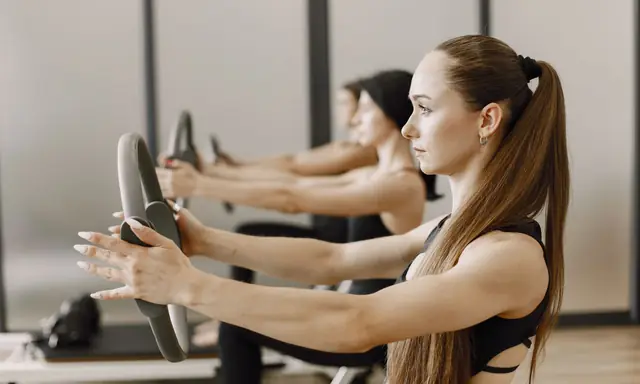 Vrouwen in een pilatesles houden een grijze pilatesring met gestrekte armen voor zich en knijpen de handvatten naar elkaar toe