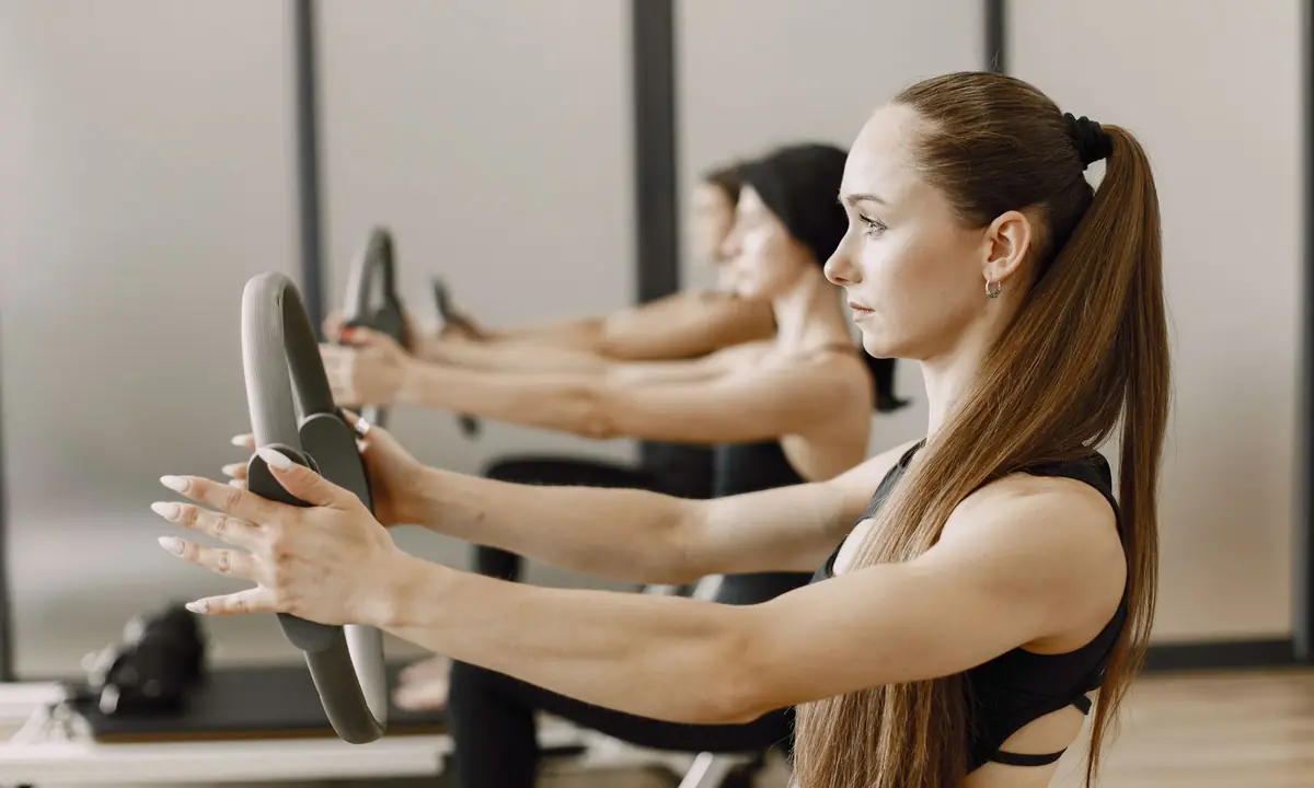 Vrouwen in een pilatesles houden een grijze pilatesring met gestrekte armen voor zich en knijpen de handvatten naar elkaar toe