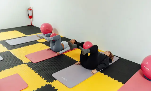 Twee vrouwen liggen op hun rug op matten met een kleine roze pilatesbal tussen hun onderbenen; rechts ligt een grote gymbal